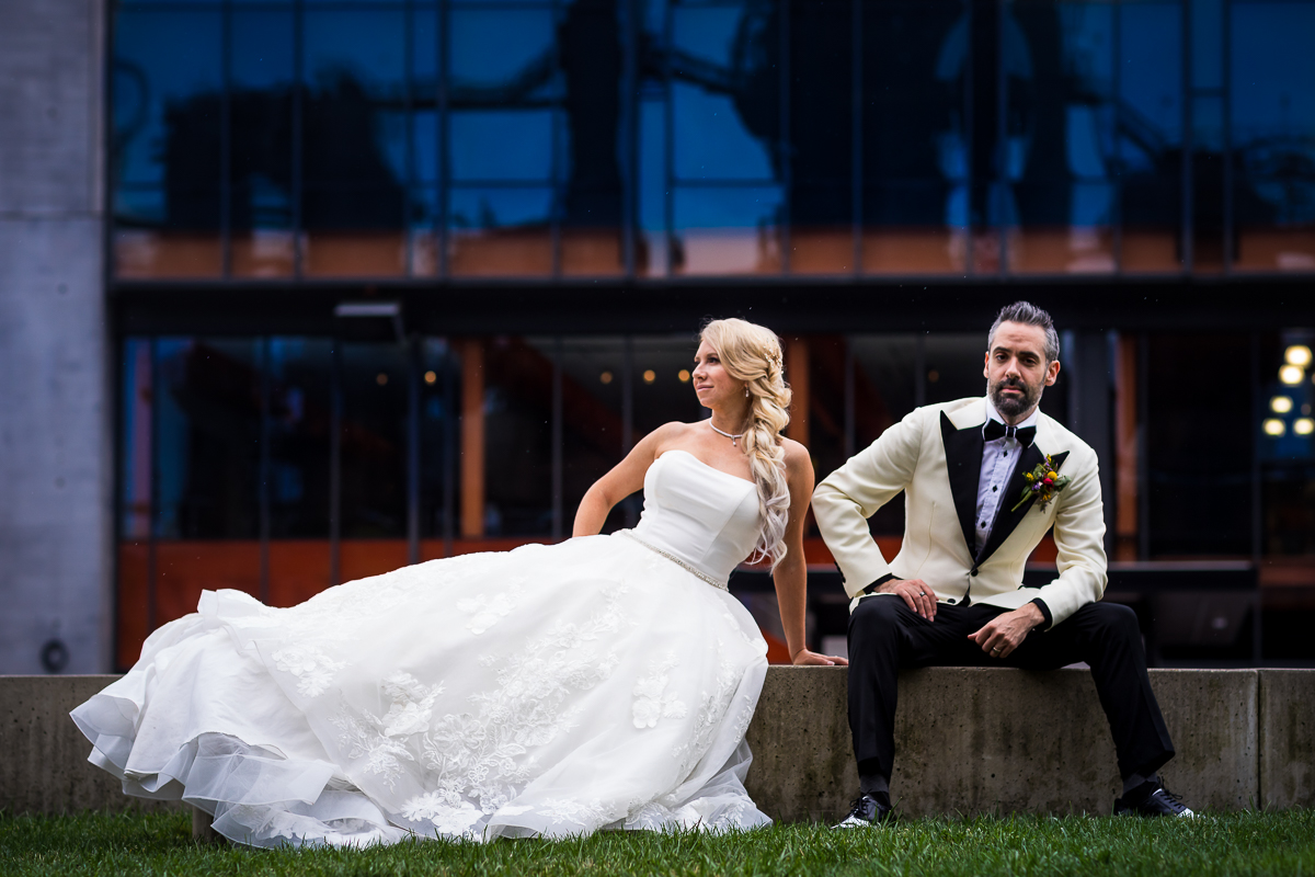 bride and groom sit on concrete bride with hand on hip and groom with hand on knee posing vogue style with steelstacks reflected in windows behind them