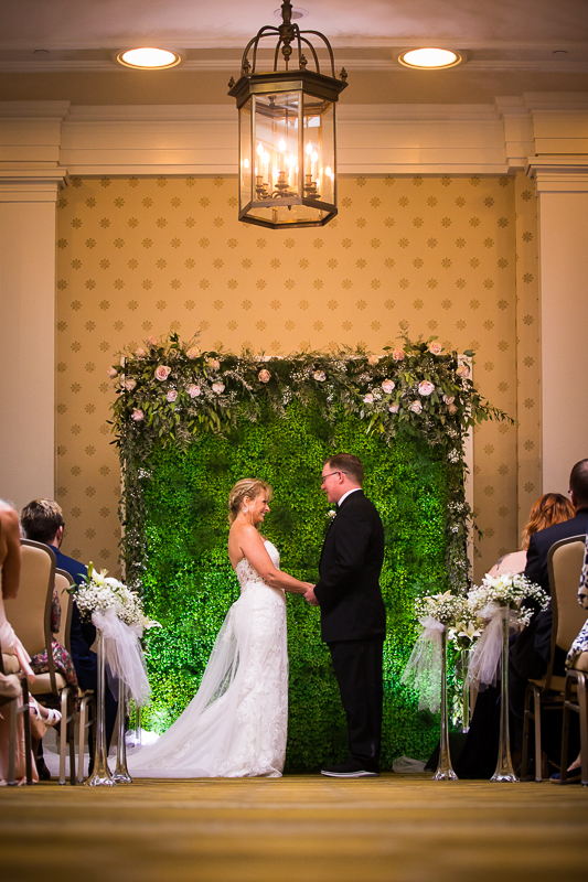 Omni Bedford Springs wedding photographer constitution hall ceremony couple stands and holds hands looking at each other before ceremony starts in front of greenery wall