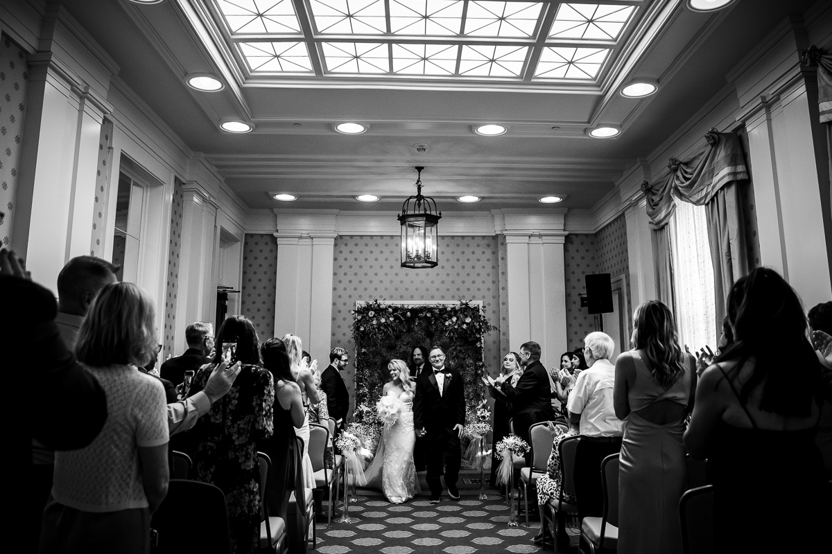 black and white phot of couple leaving ceremony after becoming husband and wife in constitution hall at Omni Bedford Springs