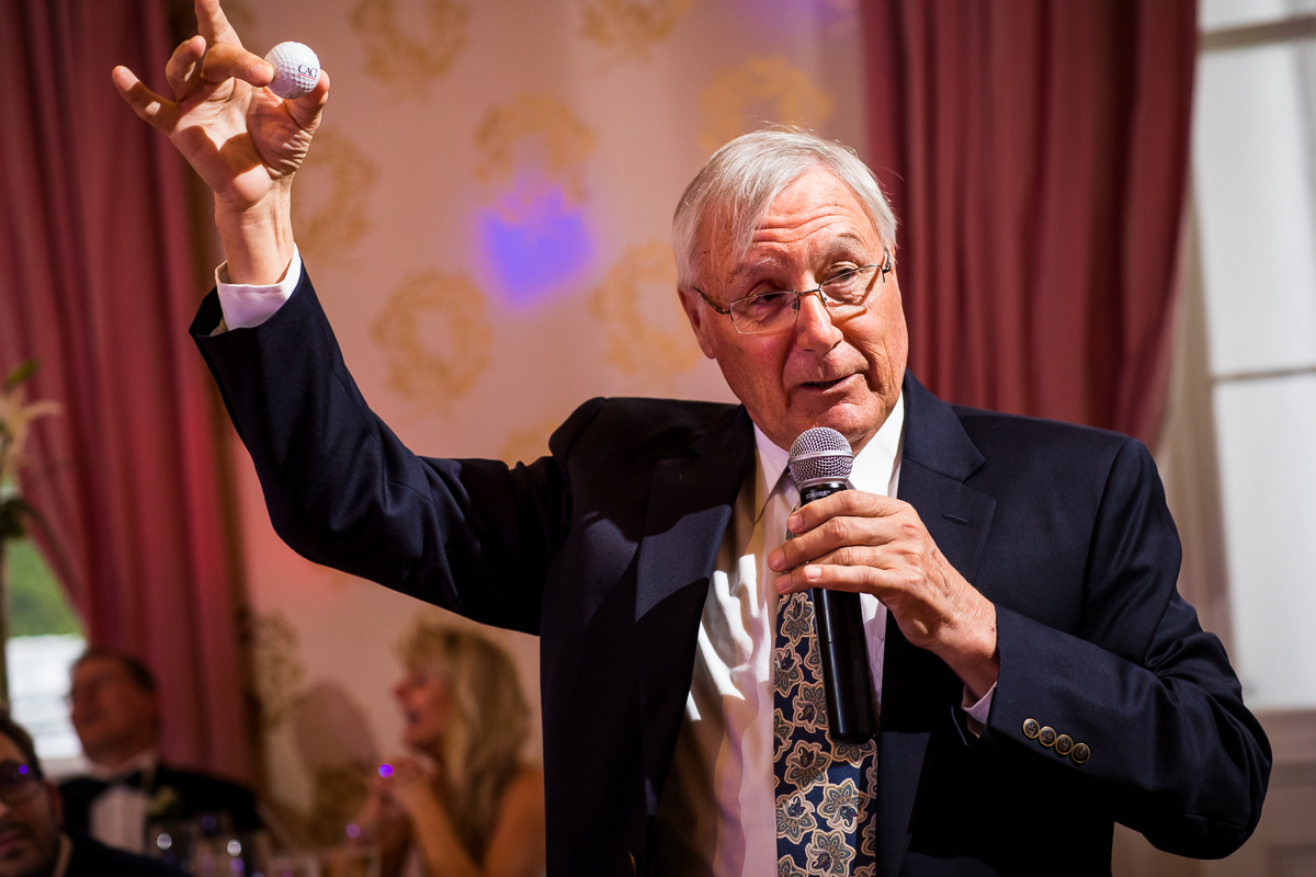 groom's uncle holds up golf ball during wedding speeches while couple laughs behind them at wedding reception central pa