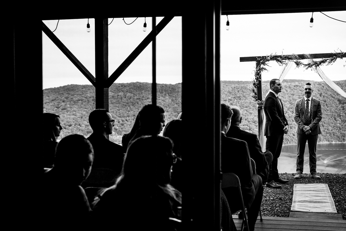 black and white photo of groom and officiant waiting under ceremony arch for bride with guests looking on 