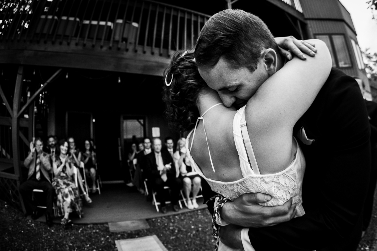 black and white photo of bride and groom hugging with guests cheering and clapping behind them