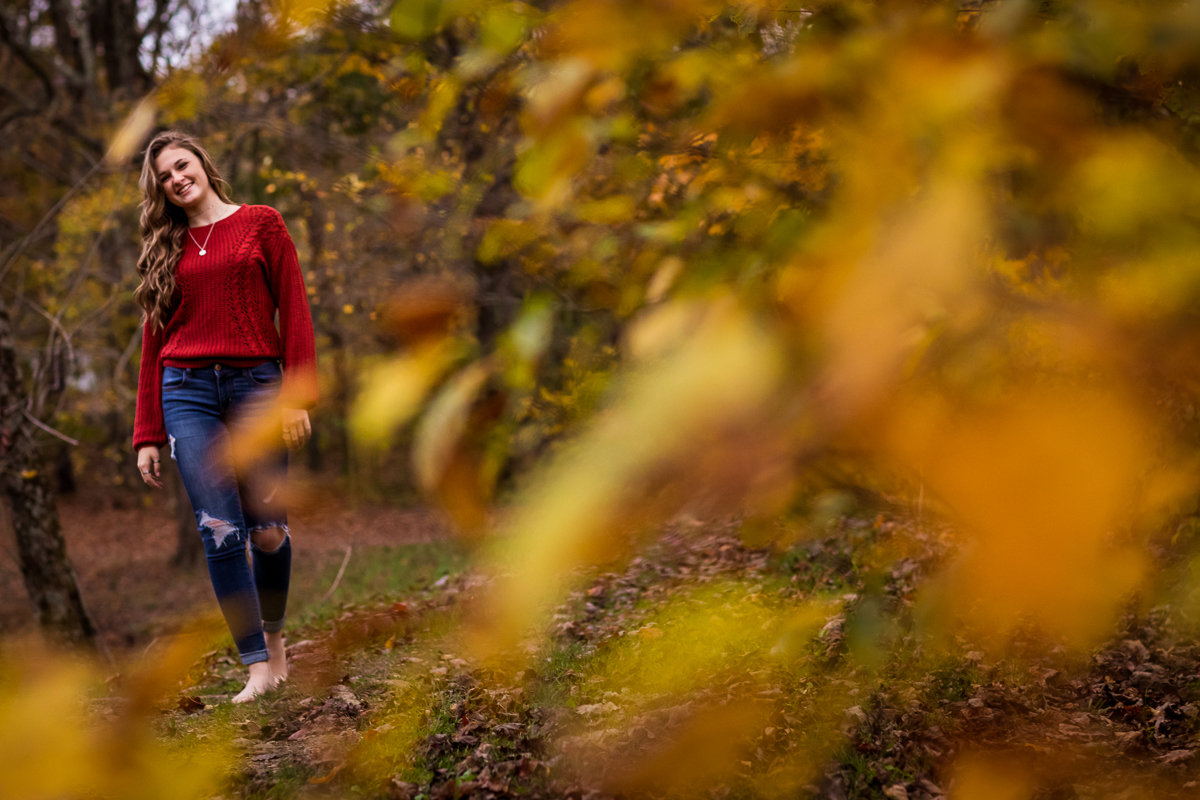 girl walking through park surrounded by colorful leaves during fall senior portrait session