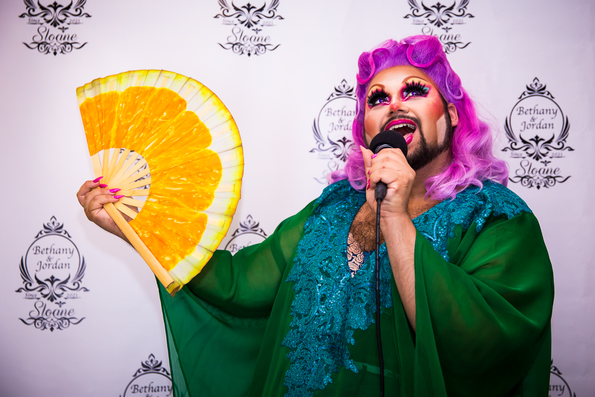 drag queen posing with orange fan and purple hair with microphone at wedding