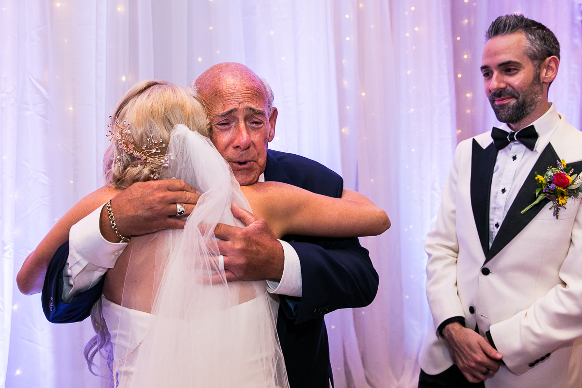 bride hugging guest as groom looks on emotional candid authentic wedding photographer