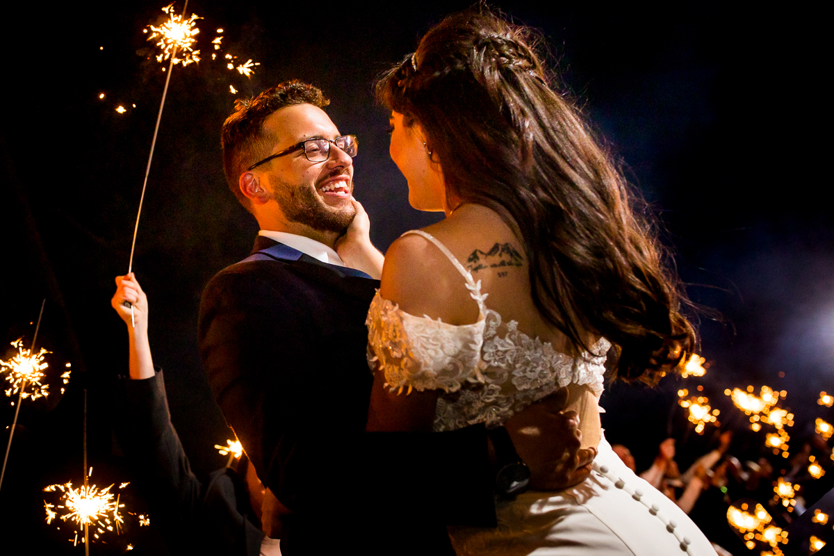creative artistic bride holding grooms face smiling at each other with sparklers behind them