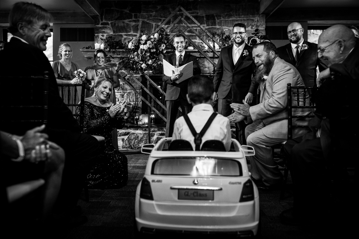 black and white photo of ring bearer driving down aisle in Mercedes with wedding guests clapping and cheering