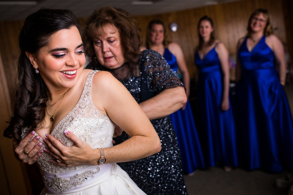 bride mom helping her get dressed while bridesmaids smile in background candid authentic real life wedding photographer 