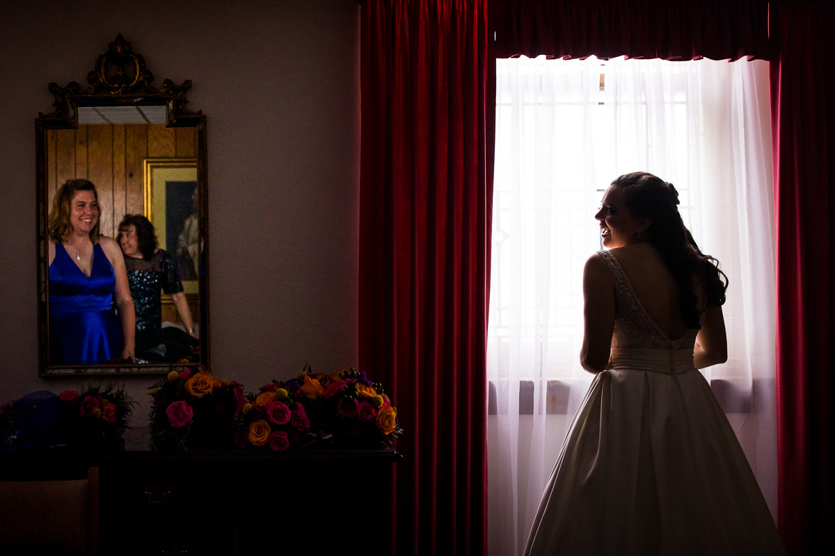 bride silhouetted against window while bridesmaids and mom reflected in mirror smiling 