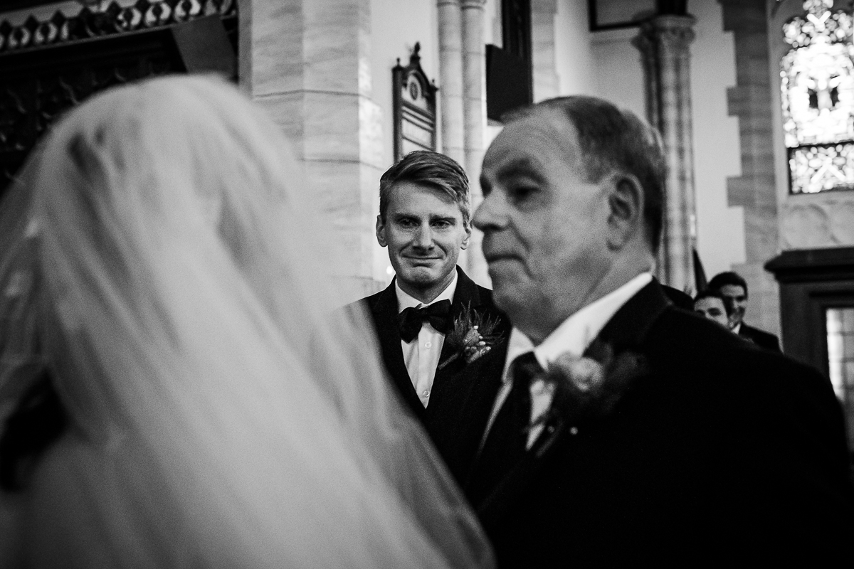 black and white photo of groom crying while seeing bride on wedding day at end of aisle