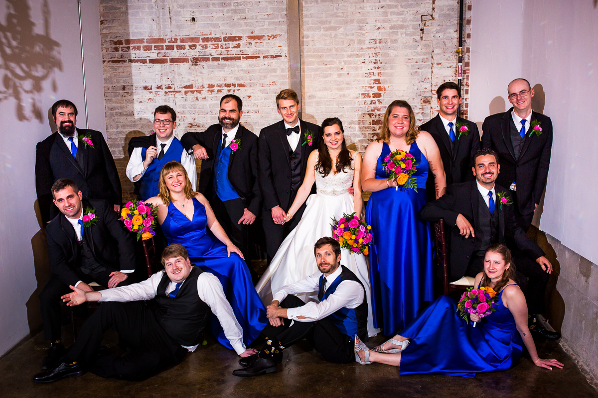 bride and groom posing with wedding party during the bond wedding in front of brick wall cobalt and bright colors