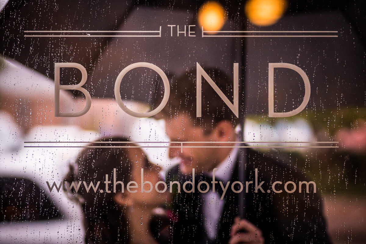bride and groom looking at each other holding umbrella reflected into the bond sign while raining