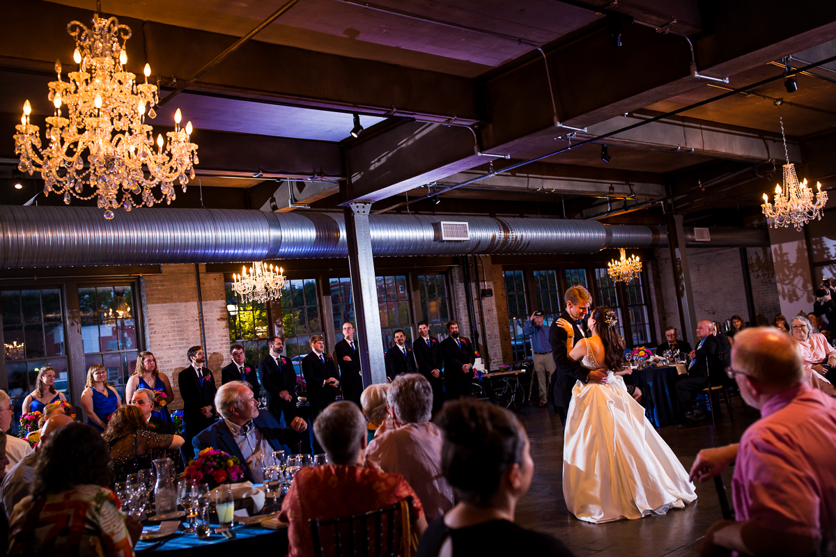 bride and groom in middle of dance floor during first dance guests smiling at them chandeliers above the bond wedding