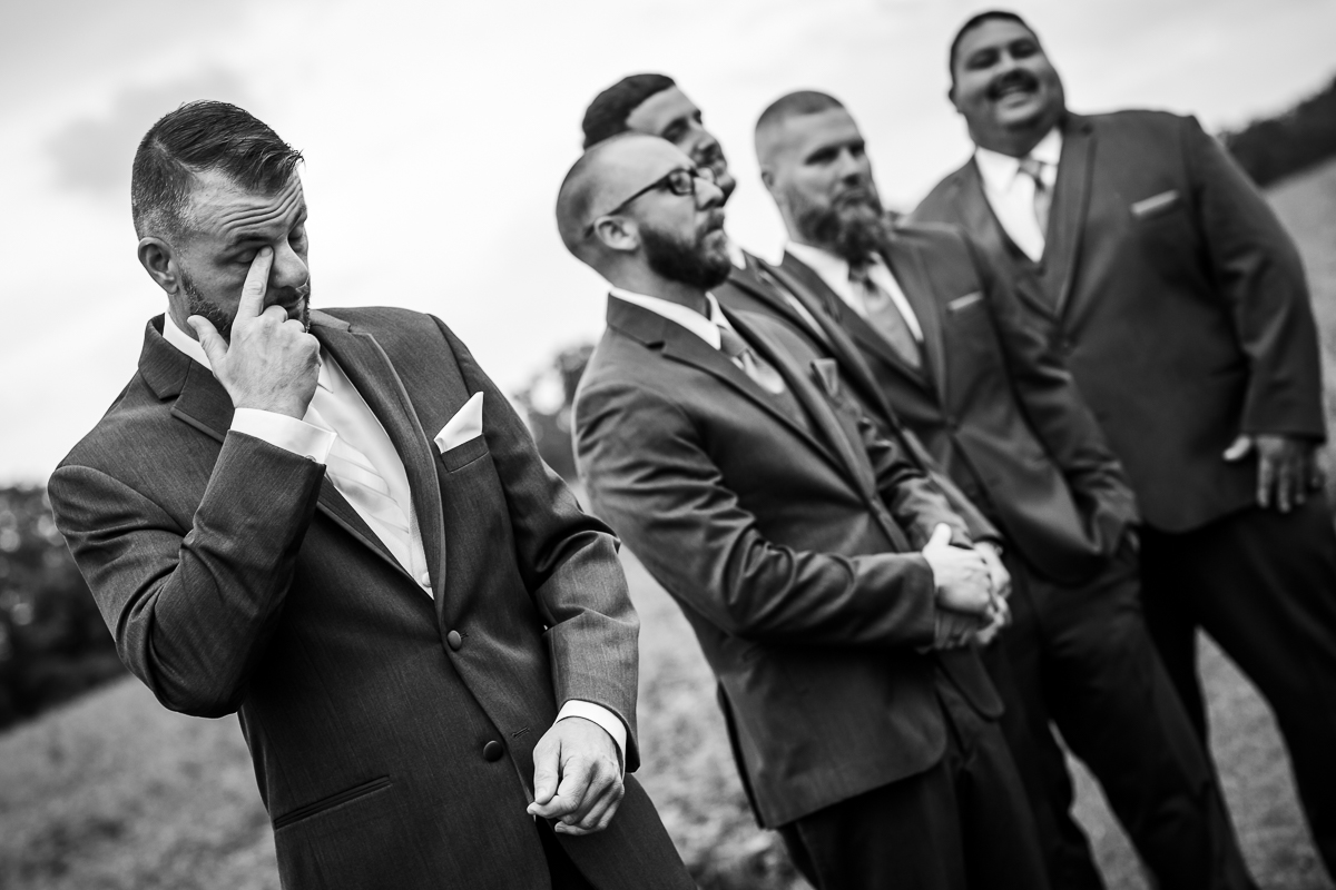 black and white photo of groom wiping tear away from eye as bride walks up to ceremony 