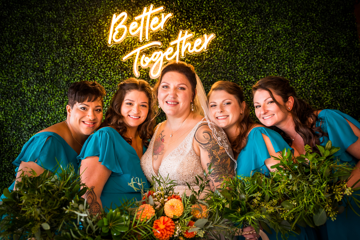 bride and bridesmaids wearing teal dresses standing in front of faux greenery wall with better together neon sign behind