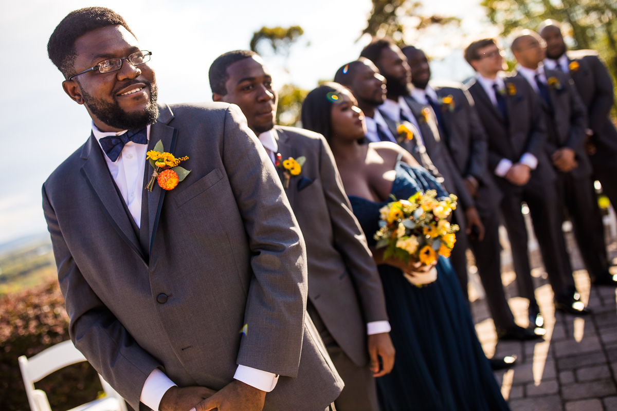 groom watching for bride as she walks down aisle smiling wedding party members stand behind