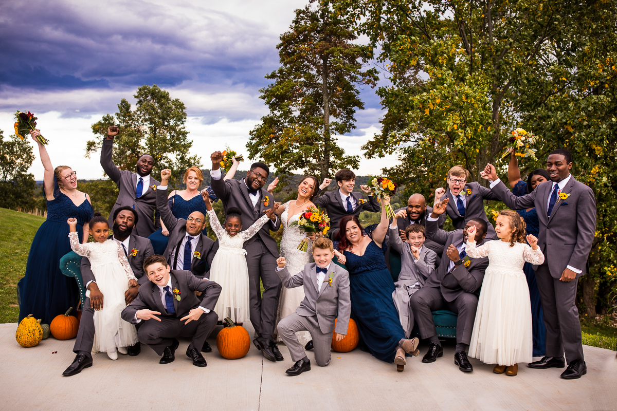 bride and groom posing with wedding party holding sunflower bouquets sitting with pumpkins 
