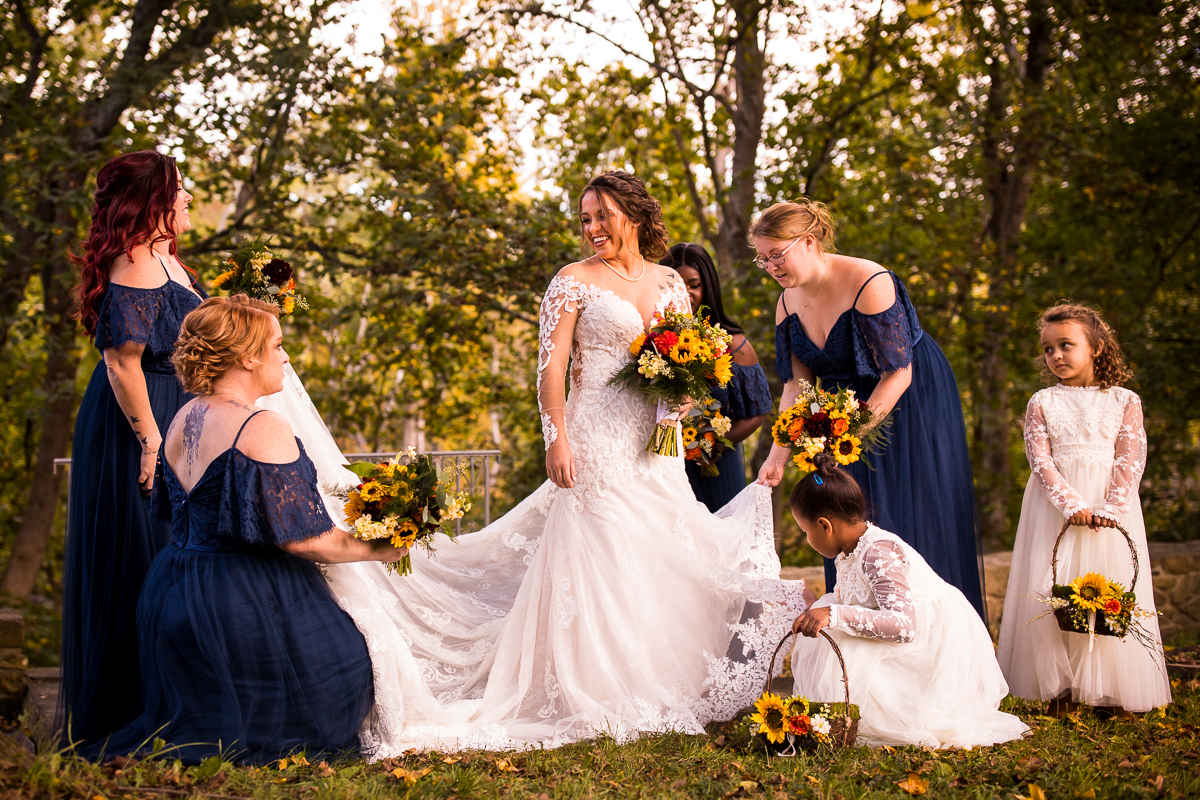 bride and wedding party members helping fluff her dress before pictures navy dresses holding colorful sunflower bouquets