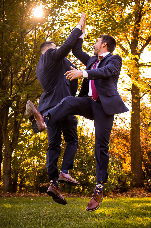 groomsmen jumping up high fiving together outside with leaves and sun shining