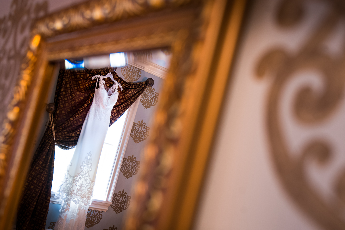 wedding dress hanging in front of window reflected into ornate gold mirror