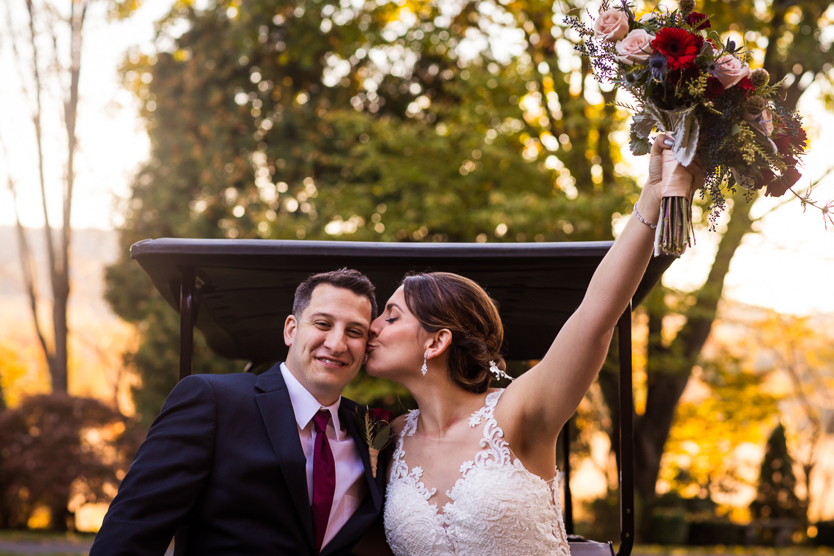 bride and groom riding on the back of golf cart during golden hour photos bride kisses groom on cheek while holding bouquet in air Linwood estate wedding photographer