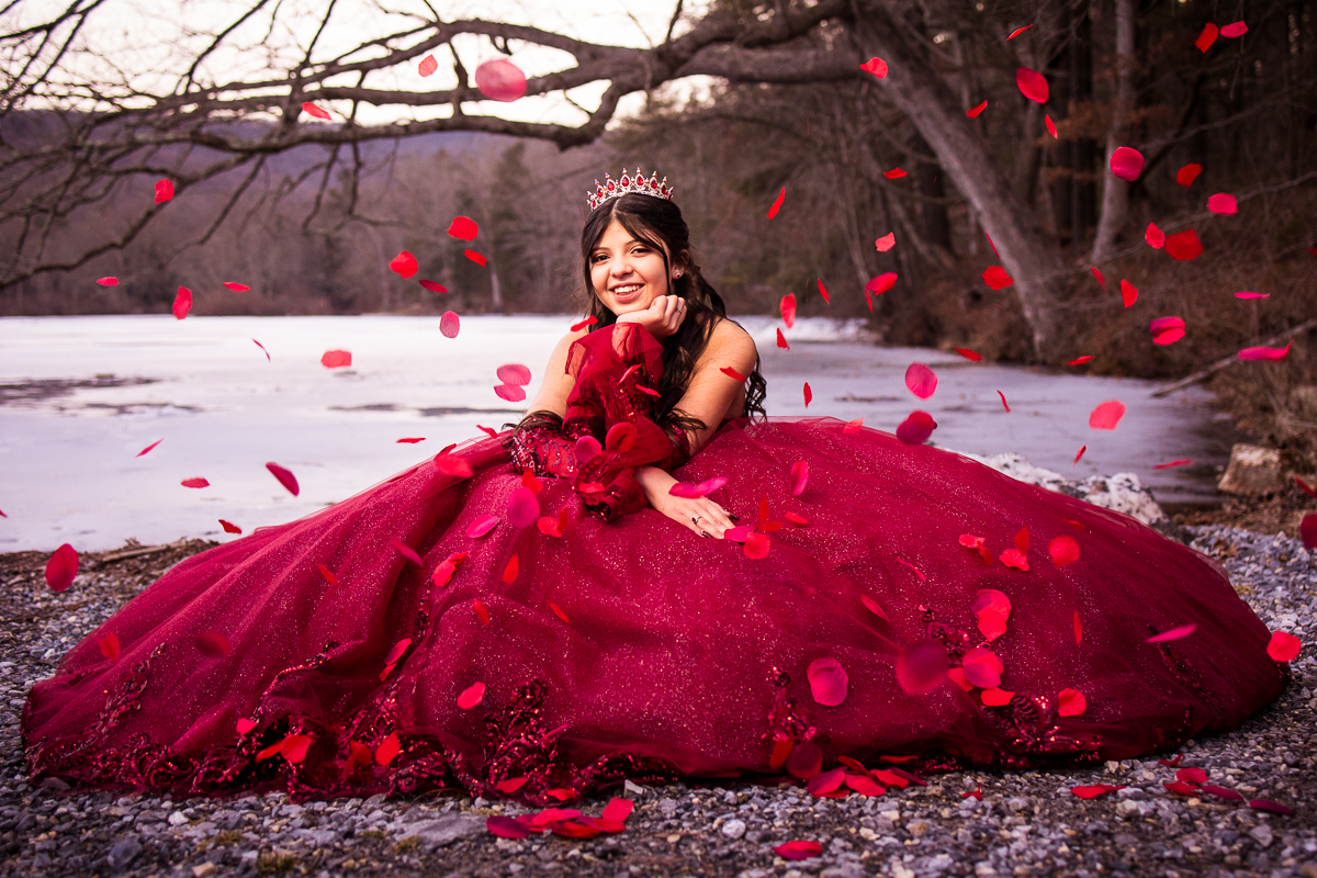 girl wearing red quinceanera dress surrounded by rose petals in the snow in forest