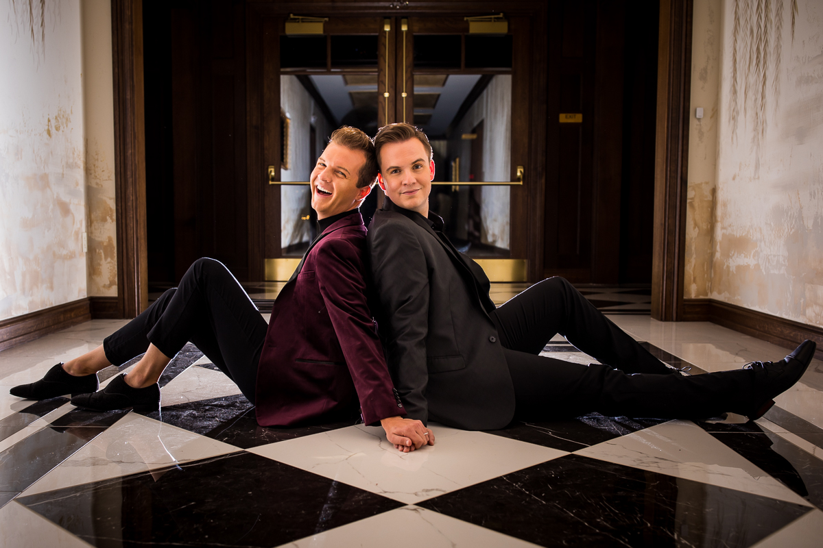 Gay couple sitting together on black and white checkered floor inside of the mansion for their engagement session with dramatic lighting