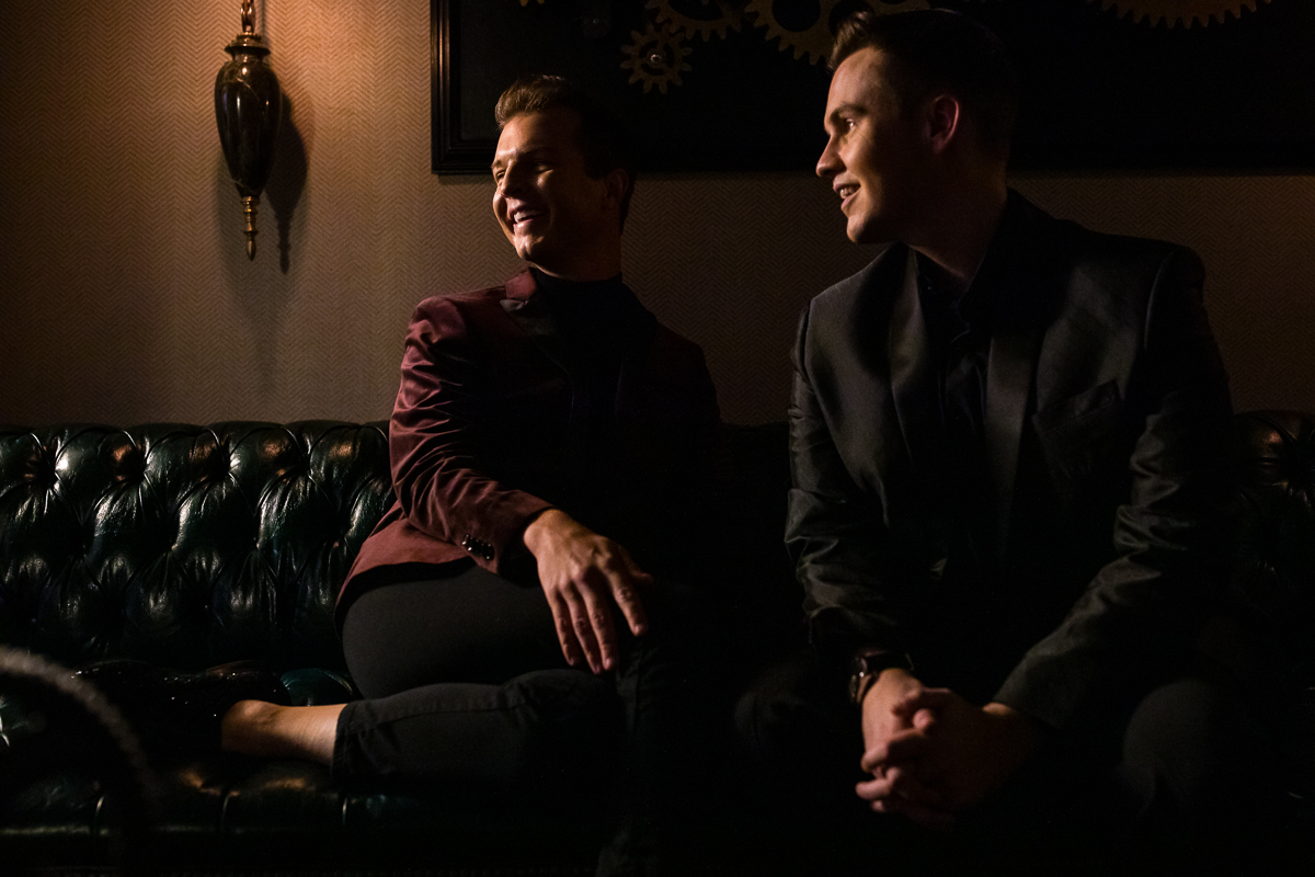 Gay couple sitting together on black couch looking away from the camera with dark, dramatic lighting for this Willows at Ashcombe Mansion engagement session