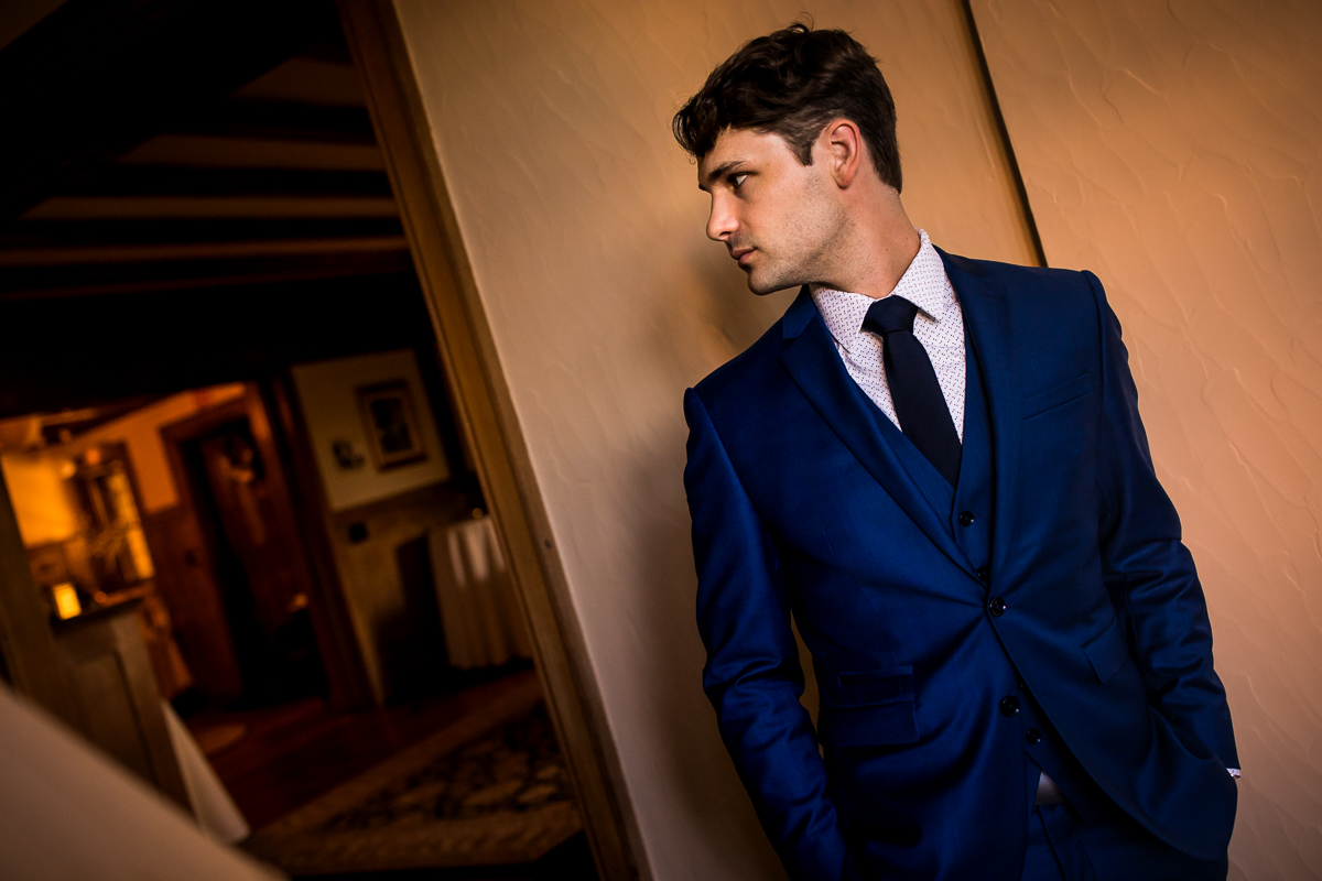 Photo of the groom standing against a light colored wall in his navy blue tux looking away from the camera