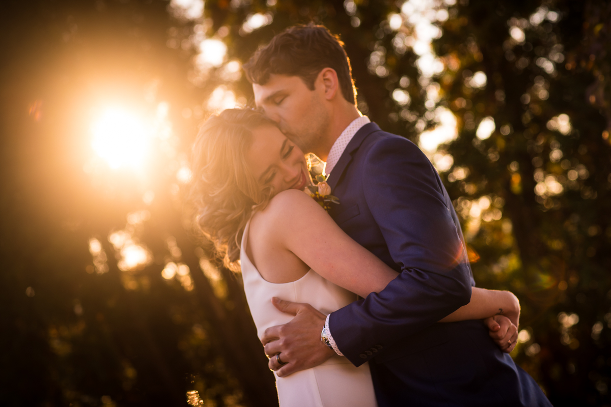 The couple is hugging each other tightly while the groom kisses the brides forehead and they are backlit by the sunset peaking through the trees for this Civic Club of Harrisburg wedding