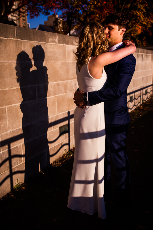The bride and groom are dancing outside with each other while their shadows are captured on the brick wall dancing behind them