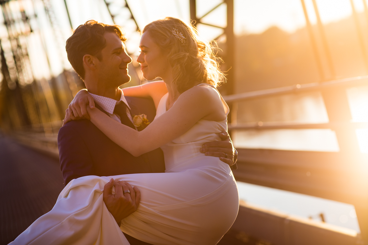 The groom is carrying the bride over a bridge in his arms and they are looking at one another smiling in this golden hour photograph