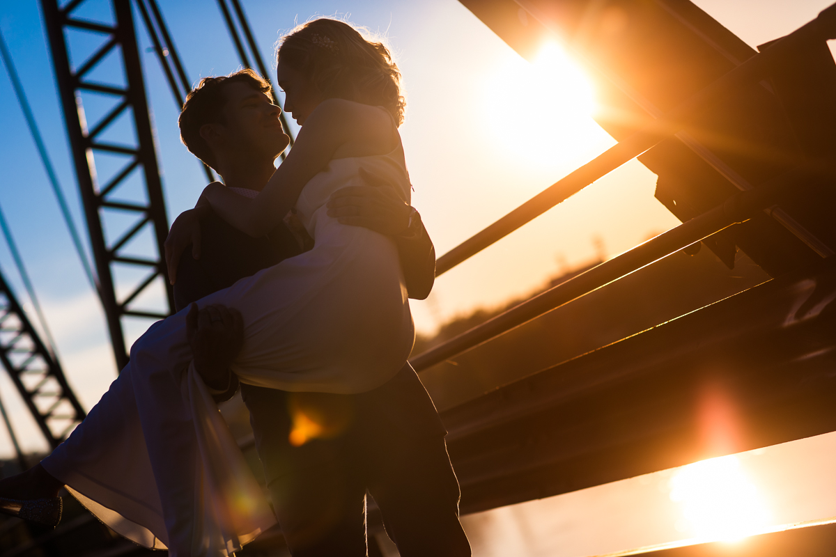 The groom is carrying the bridge down the bridge in his arm while they look towards each other in this golden hour sunset photo for this Civic Club of Harrisburg Wedding