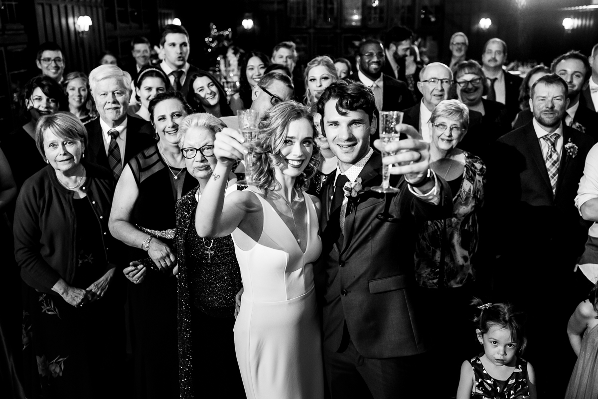 Black and white image of the couple holding up glasses toasting to the camera with their guests standing behind them as a nod to their favorite Stephen King novel for this Civic Club of Harrisburg wedding reception