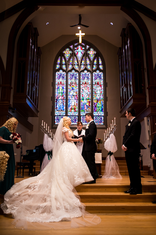 Bride and Groom holding hands and smiling at each other during their wedding ceremony in front of a colorful stained glass window inside First United Church Hershey Wedding Ceremony