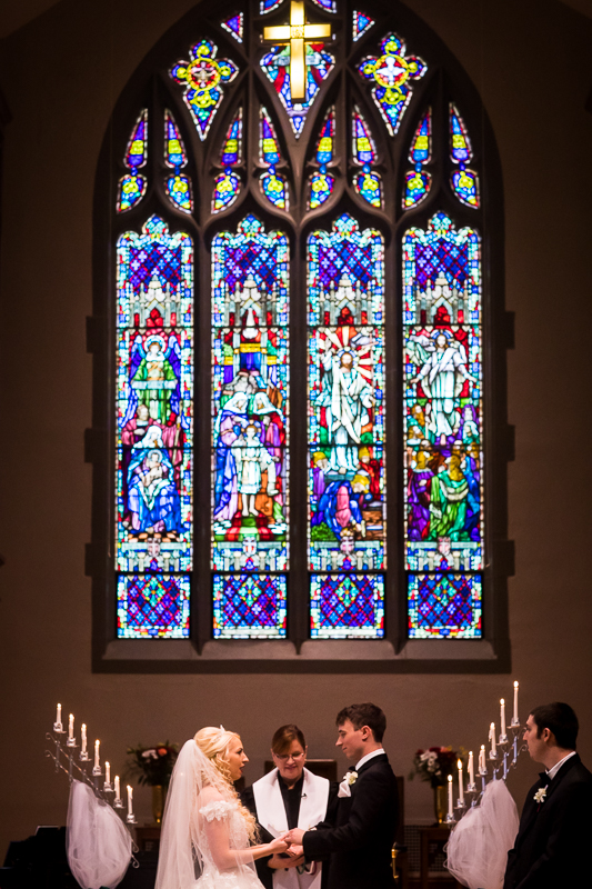 Bride and Groom holding hands looking at each other with a bright and colorful stained glass window as the focus of the photo taken inside First United Church Hershey Wedding Ceremony