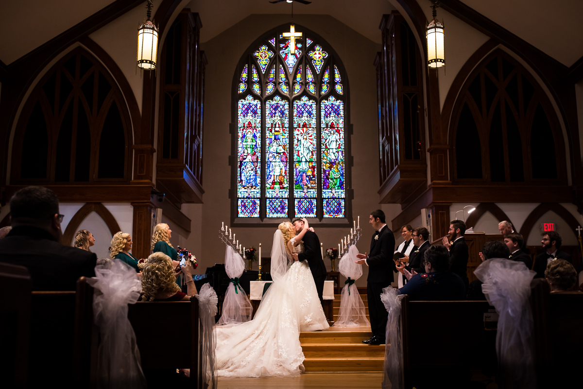 Bride and Groom kissing during their wedding ceremony inside of First United Church Hershey Wedding Ceremony with a large bright and color stained glass window in the background