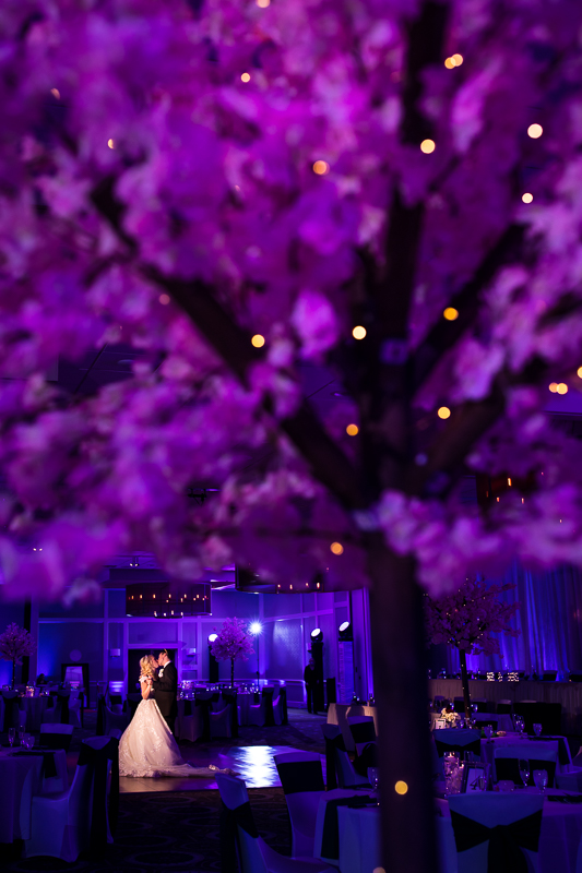 Creative and vibrant colored photo peeking through the lighted tree center pieces of the bride and groom dancing at their wedding reception inside Best Western Harrisburg