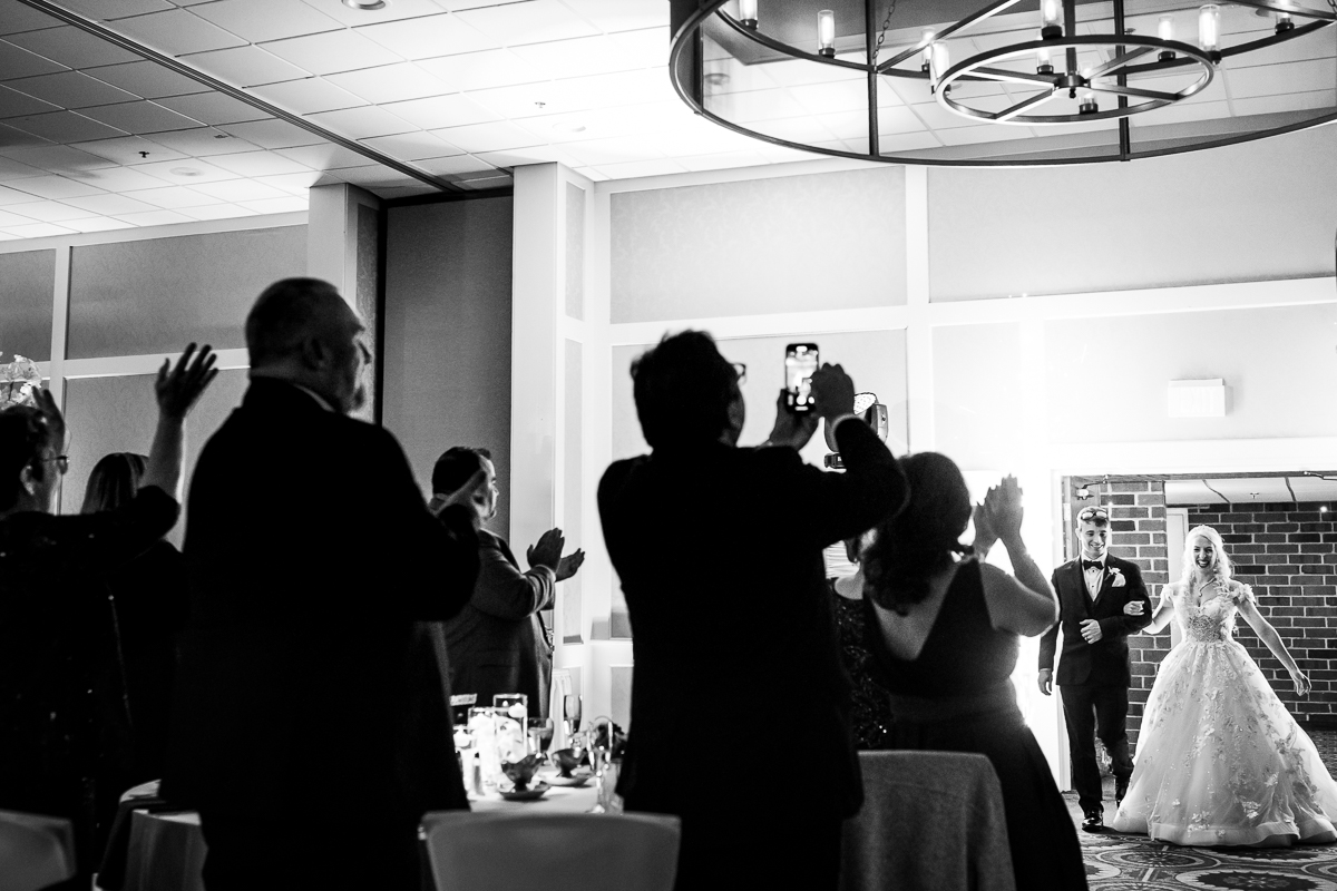 Black and white photo of the bride and groom walking in to their wedding reception with their guests standing up, clapping and taking photos of the couple walking in together