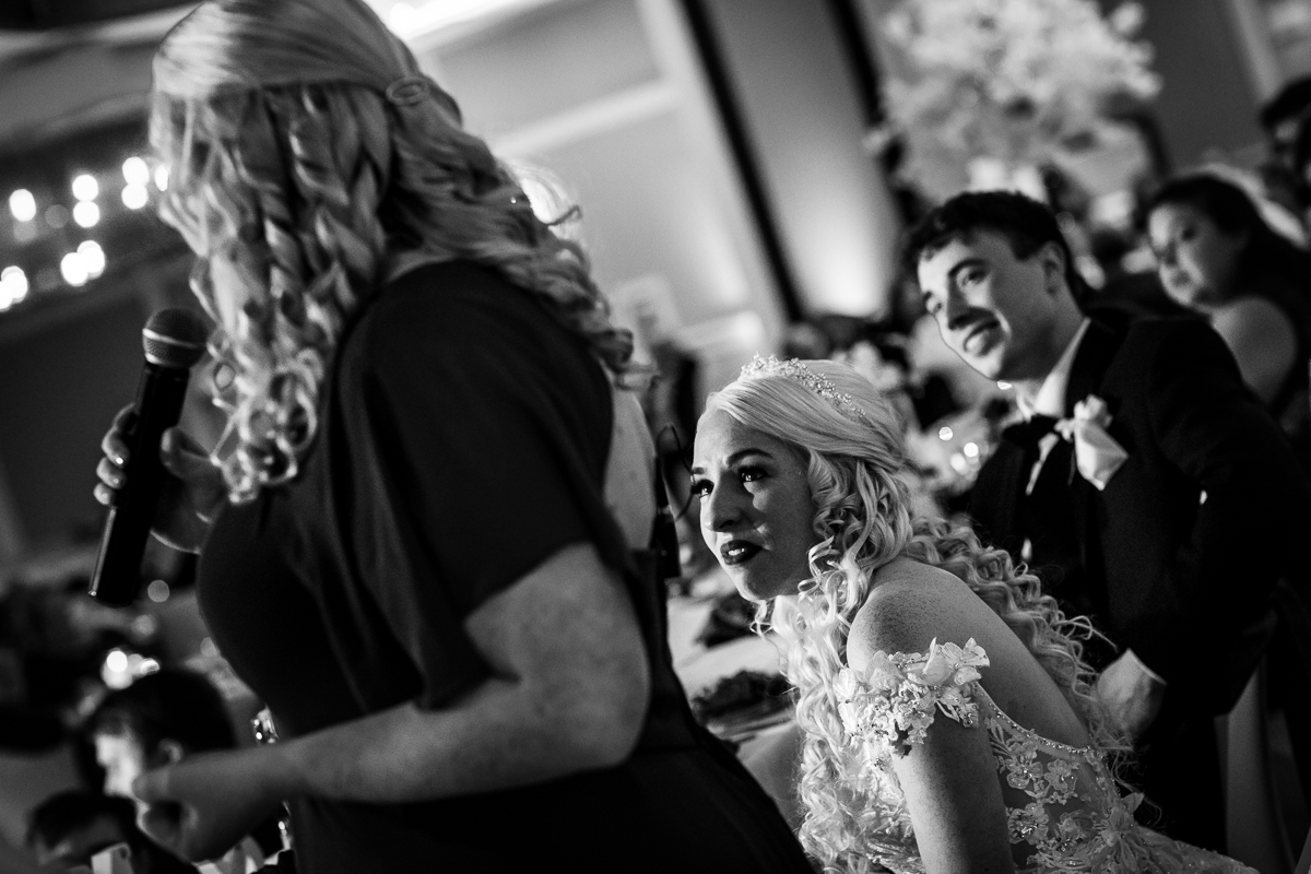 Black and white photo of the bride and groom looking at a bridesmaid giving a speech during their wedding reception 