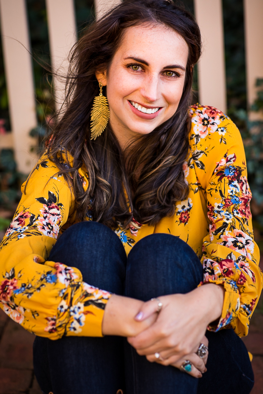 traditional portrait of travel blogger in her bright yellow floral shirt and feathered earrings hugging her legs as she smiles at the camera for this bethleham portrait photographer, rhinehart photography