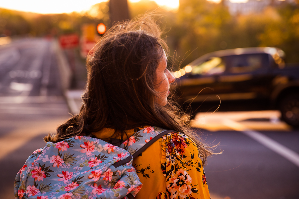 colorful image of travel blogger lauren walking across the street looking out towards the road in her yellow floral shirt and blue and pink floral backpack in this arts quest photographer image by rhinehart photography