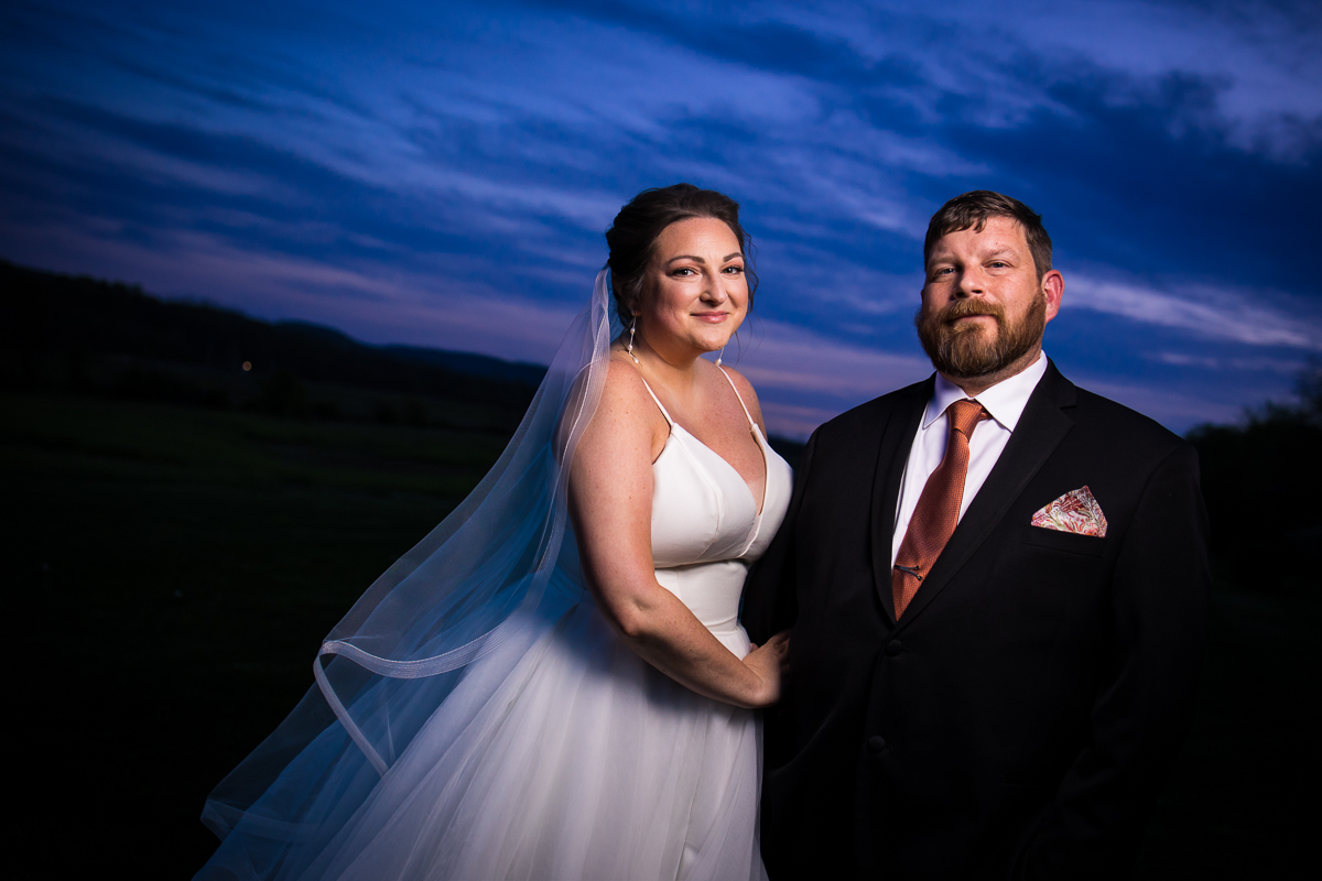 Colorful, creative image of the bride and groom smiling at the camera with a dark blue, purple sunset behind them after their Linwood estate wedding reception