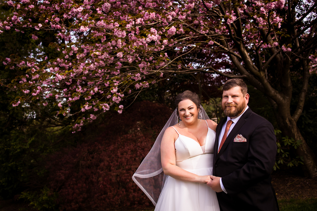 traditional portrait of the bride and groom in front of a pink blossoming tree holding hands after their wedding ceremony at linwood estate