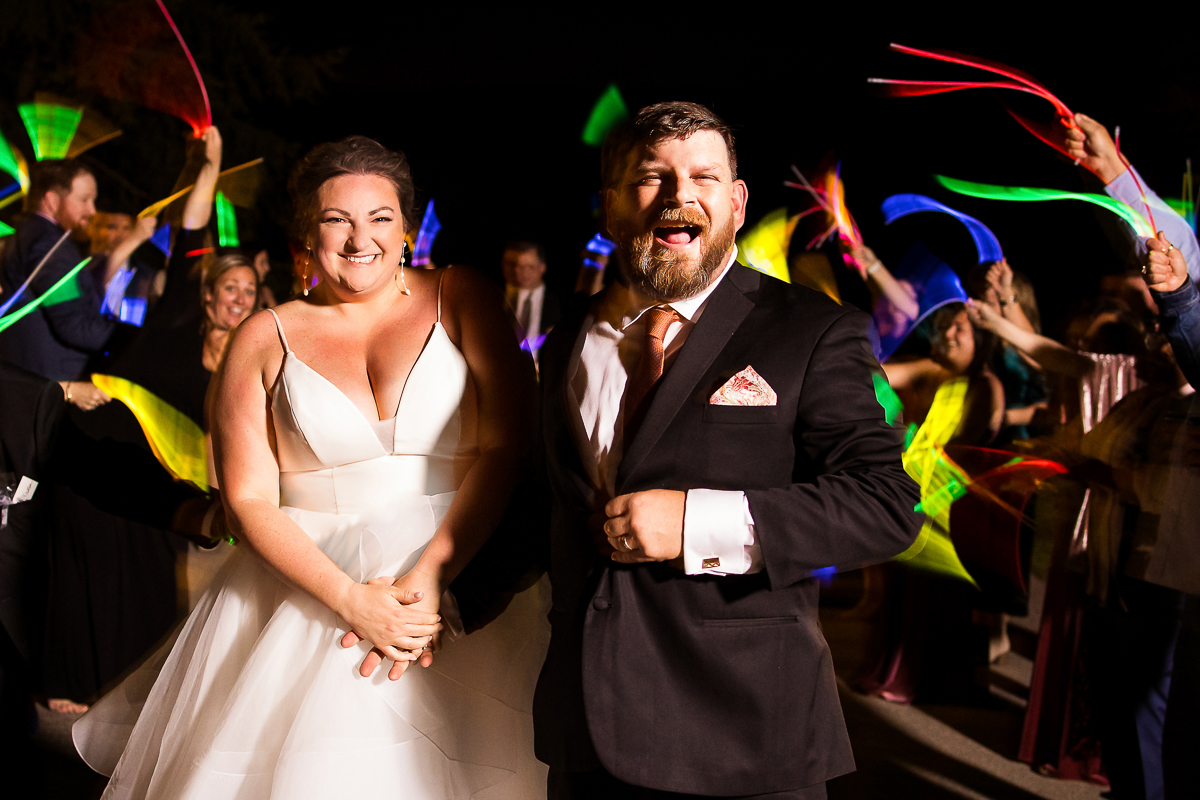 vibrant, colorful, unqiue image of the bride and groom leaving their reception with guests waving glowsticks in the background during this linwood estate wedding 