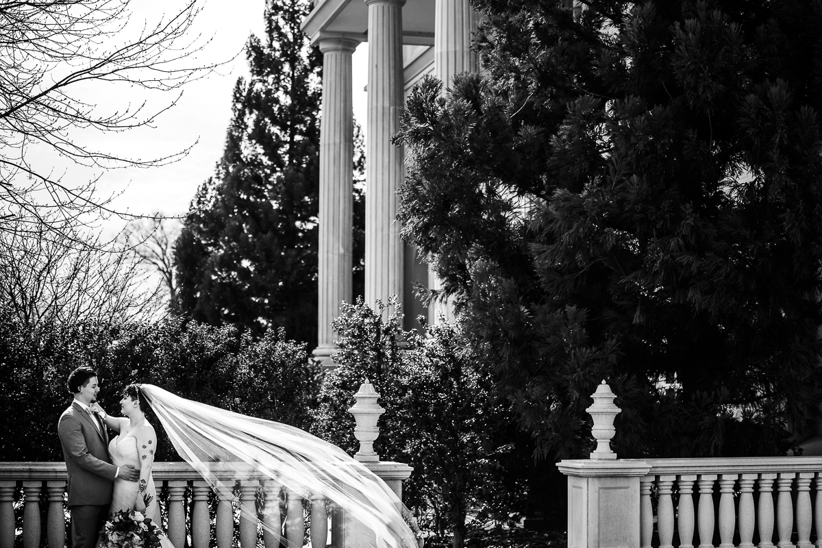 black and white image of the bride and groom hugging one another as they stand outside of the palace in new jersey as the bride's veil blows in the wind behind her