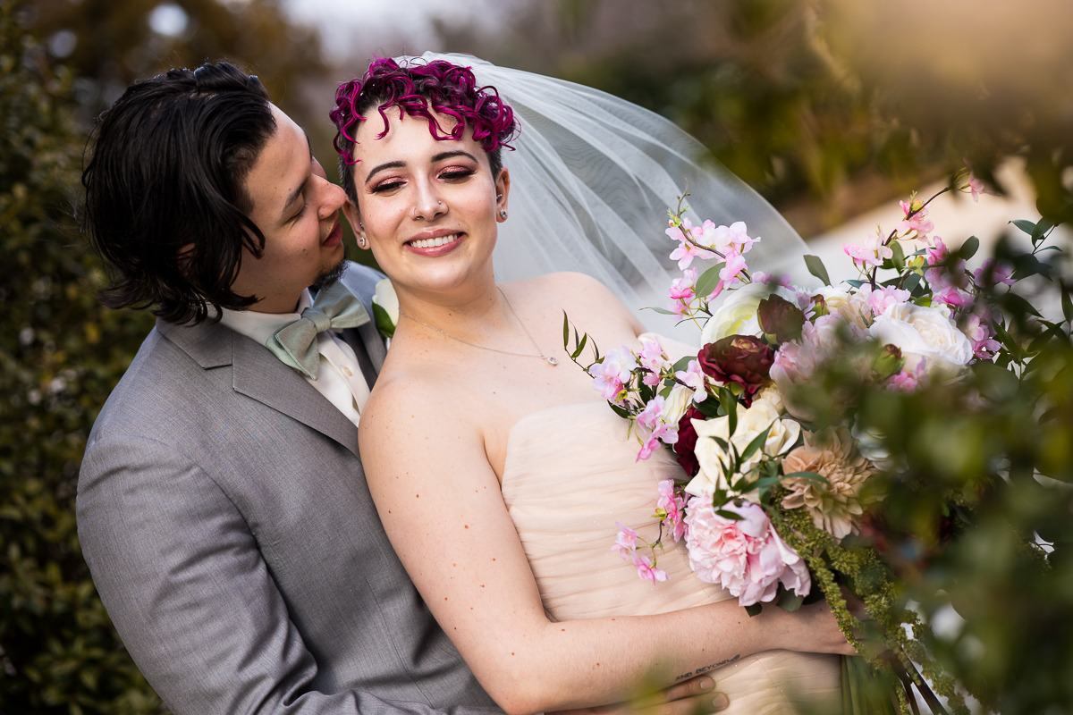 Colorful, vibrant, image of the bride smiling as the groom leans in towards her as they stand outside amongst trees before their Palace at Somerset Park Wedding