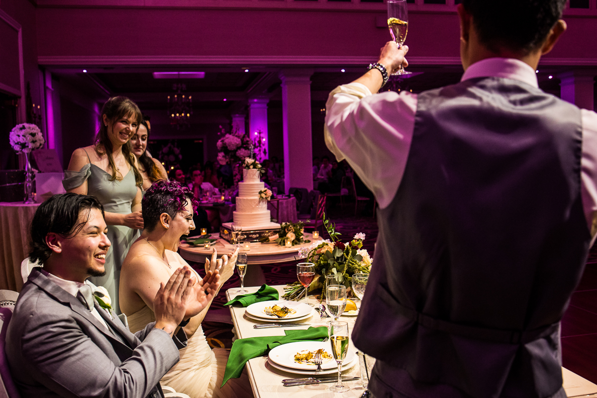 vibrant, colorful image of the wedding party giving their toasts as the bride and groom smile and clap during their wedding reception