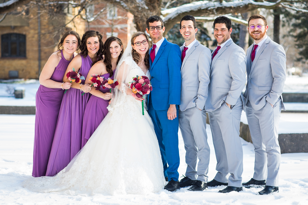 traditional portrait of the wedding party and bride and groom smiling at the camera as they are outside in the snow getting their photos taking before this Rochester NY Wedding