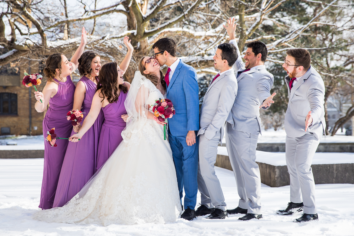 image of the groom kissing the brides cheek while the wedding party cheers around them while they stand outside in the snow before their Rochester NY Wedding